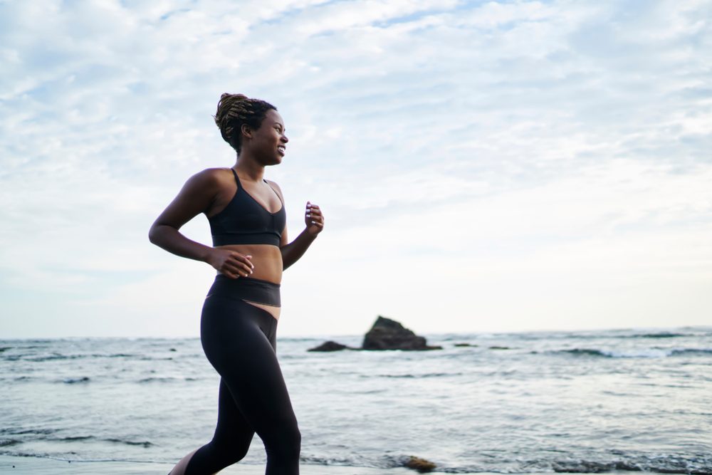 Side view of African American female jogger dressed in stylish sport clothes running along coastline washed by ocean water. Sportive fit woman with dark skin doing morning cardio training on seashore